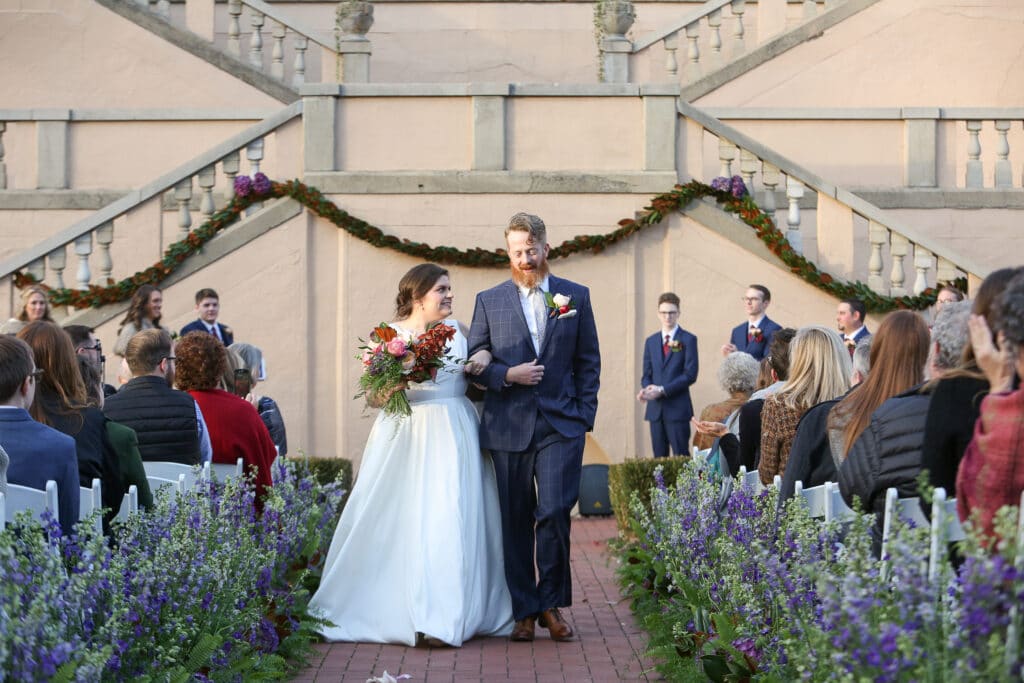 bride and groom walk down a flower-lined aisle at outdoor ceremony at fowler house mansion in lafayette, indiana