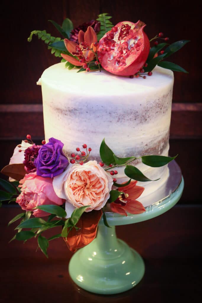three-layer wedding cake decorated with fresh flowers and pomegranates on mint glass cake stand